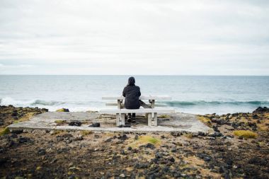 Isolated nostalgic woman siting on a bench by the sea.Pensive female thinking by the calming ocean waves.Pensive person struggling with mental problems,depression and anxiety.Rethinking and reflecting clipart