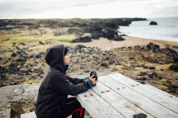 在寒冷多云的天气下 在户外露营场所的热水瓶杯中饮用热茶或咖啡饮料 在冰岛旅行的妇女穿着防水 防风夹克和保暖 — 图库照片