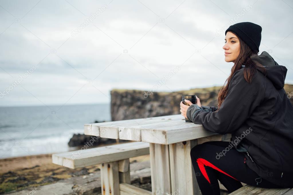 Drinking hot tea or coffee beverage in thermos flask cup in camping site outdoor on a cold cloudy day.Woman traveling around Iceland.Wearing waterproof,windproof jacket and keeping warm.Alone