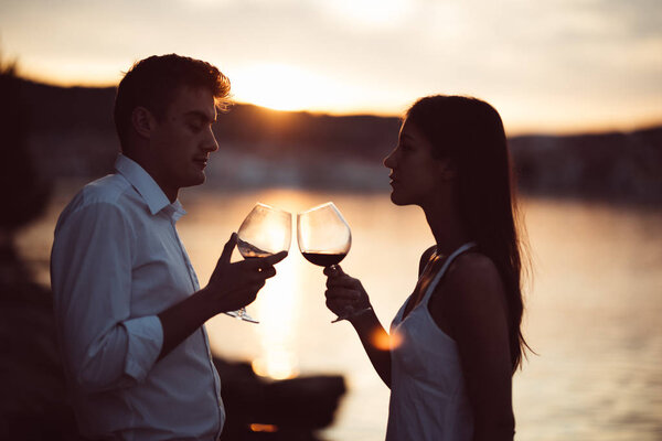 Two young people at the beach,making a toast for special occasion.Looking at the sun,enjoying the view with two glasses of wine.Drinking red wine.Celebration of anniversary.Birthday surprise.Honeymoon