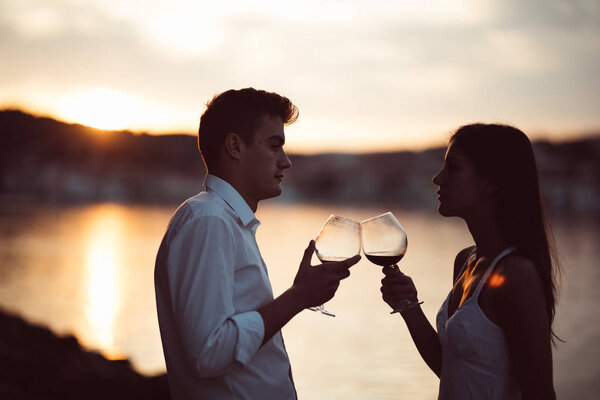 Two young people at the beach,making a toast for special occasion.Looking at the sun,enjoying the view with two glasses of wine.Drinking red wine.Celebration of anniversary.Birthday surprise.Honeymoon