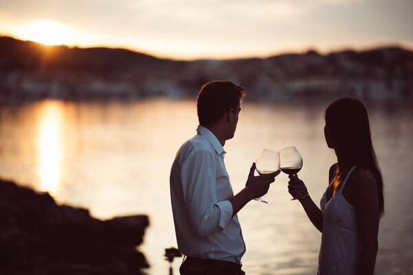 Two young people at the beach,making a toast for special occasion.Looking at the sun,enjoying the view with two glasses of wine.Drinking red wine.Celebration of anniversary.Birthday surprise.Honeymoon