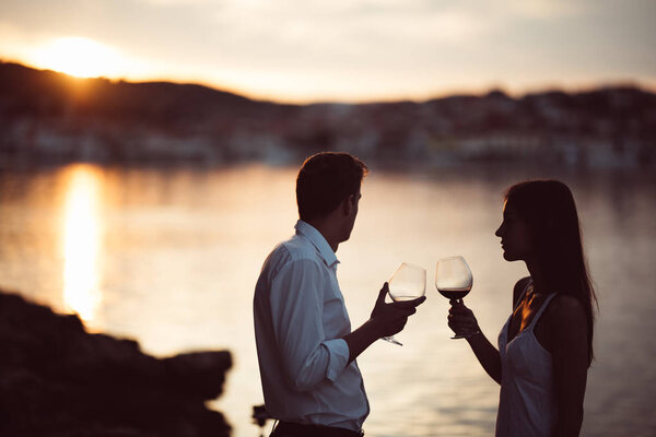 Two young people at the beach,making a toast for special occasion.Looking at the sun,enjoying the view with two glasses of wine.Drinking red wine.Celebration of anniversary.Birthday surprise.Honeymoon