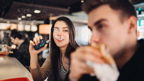 Young woman using electronic cigarette to smoke in public places.Smoke restriction,smoking ban.Using vaping device with flavoured liquid.E-juice vaping.Smoking habit,nicotine addict,tobacco industry