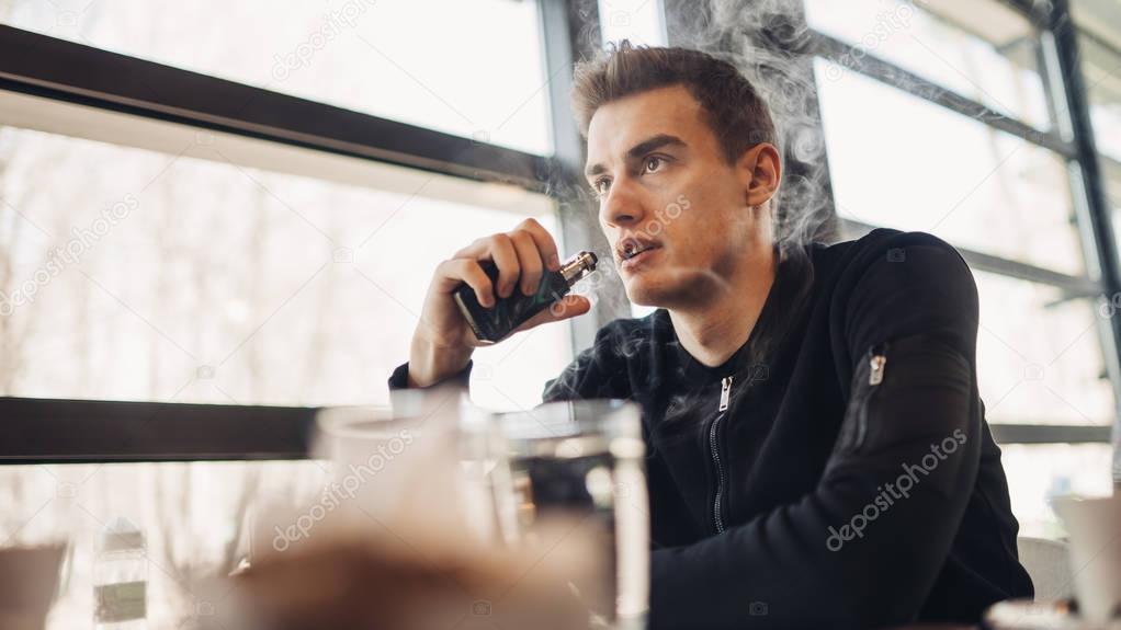 Young man vaping in closed public space.Smoking electronic cigarette in cafe.Nicotine addiction.Way to quit smoking,old habit.Vaping aroma,urban man using e-cigarette on places smoking is not allowed