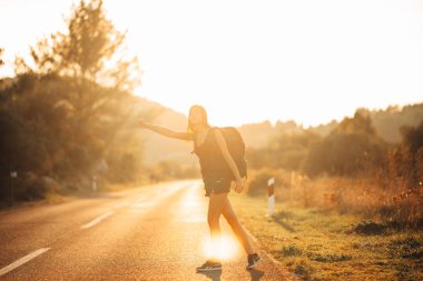 Genç, sırt çantalı, maceraperest bir kadın yolda otostop çekiyor. Başparmağıyla bir arabanın tepesinde. Seyahat tarzı. Düşük bütçeli seyahat.