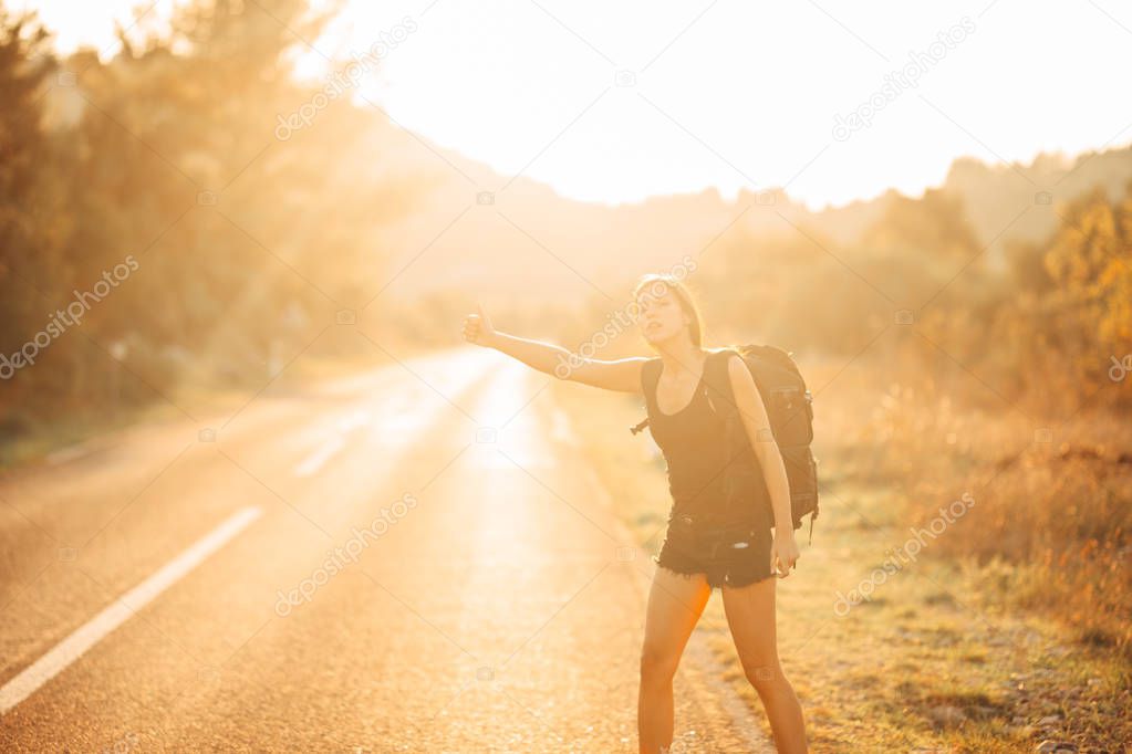 Young backpacking adventurous woman hitchhiking on the road.Stopping a car with a thumb.Travel lifestyle.Low budget traveling.Adventurous active vacations.Hitchhiking tourism concept.Backpacker