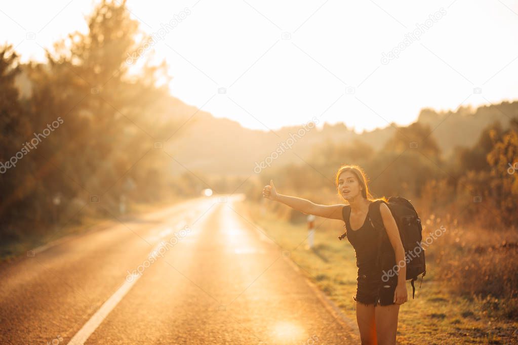 Young backpacking adventurous woman hitchhiking on the road.Stopping a car with a thumb.Travel lifestyle.Low budget traveling.Adventurous active vacations.Hitchhiking tourism concept.Backpacker