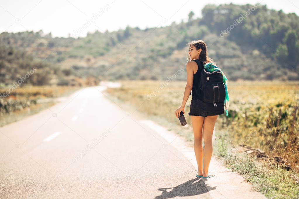 Young backpacking adventurous woman hitchhiking on the road.Traveling backpacks volume,packing essentials.Travel lifestyle.Low budget traveling.Adventurous active vacations.Hitchhiking tourism concept