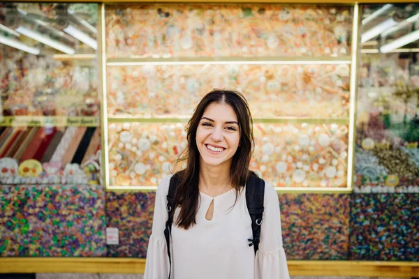 Young smiling woman laughing to camera in front of candy store.Visiting historical place.Traveling on a budget.Backpacker student roadtrip.Urban travel influencer girl