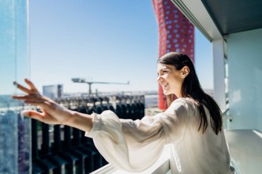 Güneşli bir günde manzaranın keyfini çıkaran genç bir kadın, sanayi / iş alanındaki apartmanın balkonunda bir pencere açıyor. Gökdelen atölyesinde yaşayan pozitif genç bir kadın. Gün için hazır. İş gezisi otel odası.