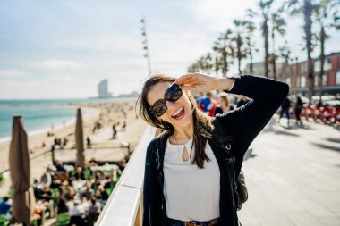 Cheerful woman enjoying sunny day at Barceloneta beach in Barcelona,Catalonia,Spain.Making funny face at camera.Energetic woman on vacation at Costa del Sol in Spain.Young backpacker clipart