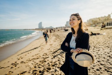 Gülümseyen neşeli kadın turist sahilde keyif çatıyor. Hasır şapkalı neşeli bayan turist, ilkbaharda Barcelona 'da kumsalda saçlarını dalgalandırıyor. Barceloneta sahilinde kadın gezgin.