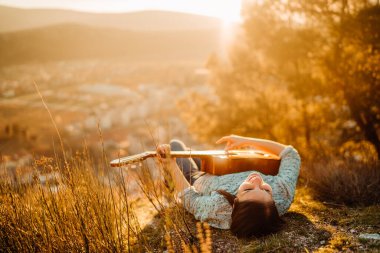 Young guitarist playing acoustic guitar and looking to sunset.Searching inspiration.Music creator.New artist in good mood.Musical talent.Smiling young woman singing and playing acoustic guitar. clipart