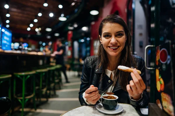 体验到的本土食物 微笑的年轻女子在传统的西班牙风味的咖啡店里 吃着用热巧克力酱汁蘸糖的传统西班牙卷心菜 — 图库照片