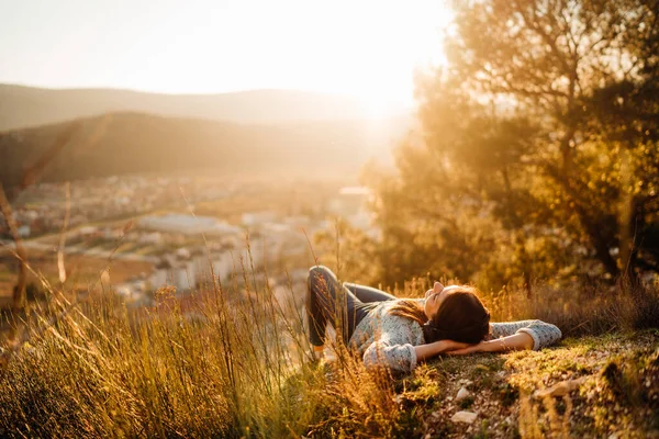 顔に太陽を楽しむ山の上に緑の芝生の牧草地に寝そべっている無料の幸せな女性 自然の夕日を楽しんでいます — ストック写真