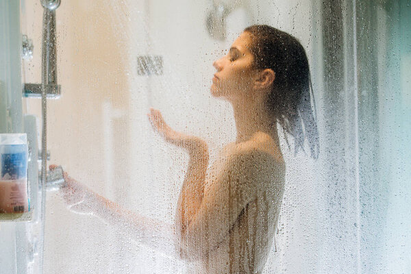 Morning shower.Taking rejuvenating cold shower.Self care moment.Everyday personal hygiene.Unfocused woman showering in glass shower with strong pressure water stream.Focus on drops