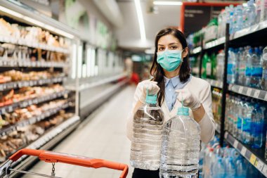 Koronavirüs salgınının ortasında marketten alışveriş yapan maskeli bir kadın. COVID-19 gıda alışverişi. Panik satın alma, stoklama. Su kıtlığı.
