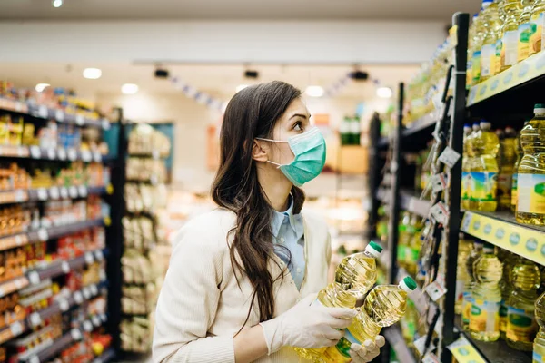 Woman with mask safely shopping for groceries amid the coronavirus pandemic in stocked grocery store.COVID-19 food buying in supermarket.Panic buying,stockpiling.Staples shortage.Cooking vegetable oil