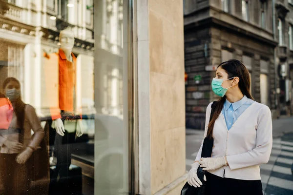 Woman with mask looking at a closed fashion clothes storefront.Clothing shopping during coronavirus outbreak shutdown.COVID-19 quarantine apparel retail store closures.Small business loss concept.