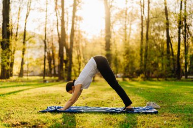 Maskeli bir kadın sadece doğada meditasyon yapıyor ve yoga yapıyor. Sosyal mesafeler ve aktif sağlıklı yaşam tarzı. Farkındalık meditasyonu. Karantina egzersizi. Aşağı doğru köpek pozisyonu.