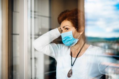 Evde oturan yaşlı üzgün kadın camdan dışarı bakıyor. Coronavirus COVID-19 salgını salgını salgını tehlikesi. Sosyalleşme kısıtlaması etkinliği.