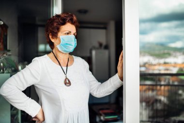 Balkon camında maske takan yaşlı bir kadın evde. Coronavirus COVID-19 hastalık salgını enfeksiyonu riski. Sosyalleşme kısıtlama etkinliği. Karantina akıl sağlığı etkisi