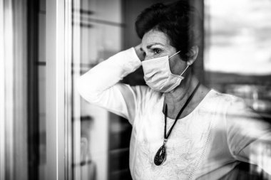 Evde oturan yaşlı üzgün kadın camdan dışarı bakıyor. Coronavirus COVID-19 salgını salgını salgını tehlikesi. Sosyalleşme kısıtlaması etkinliği.