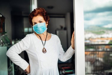 Balkon camında maske takan yaşlı bir kadın evde. Coronavirus COVID-19 hastalık salgını enfeksiyonu riski. Sosyalleşme kısıtlama etkinliği. Karantina akıl sağlığı etkisi