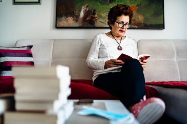Evde kitap okuyan yaşlı bir kadın. Coronavirus COVID-19 salgını enfeksiyonu riski. Sosyalleşme kısıtlaması etkinliği. Karantina zihinsel sağlık etkisi