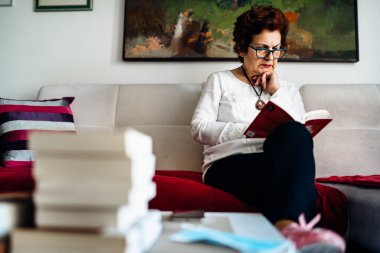 Evde kitap okuyan yaşlı bir kadın. Coronavirus COVID-19 salgını enfeksiyonu riski. Sosyalleşme kısıtlaması etkinliği. Karantina zihinsel sağlık etkisi