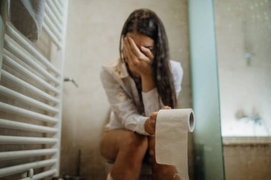 Tuvalette elinde tuvalet kağıdı olan üzgün bir kadın, karın ağrısı çekiyor. Sindirim ve üreme sisteminde kadın sağlığı sorunu. Kabızlık / regl krampları. Glüten / laktoz intolerans semptomları.