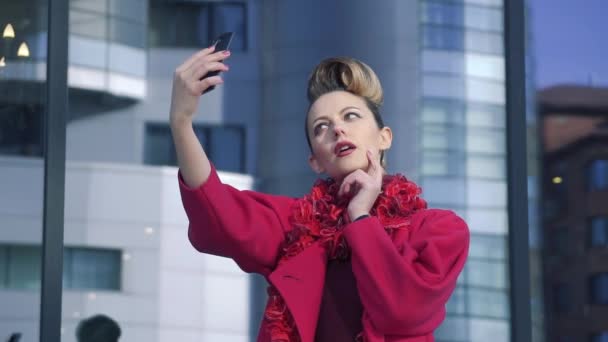 Självsäkra kvinnan med en vacker ovanlig frisörsal gör selfie våren staden. Flicka på bakgrund av modern arkitektur. Slow Motion — Stockvideo
