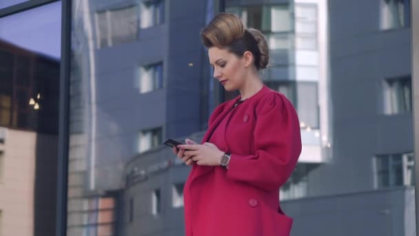 美しい珍しい髪型と深紅のコートに自信を持って女性は、春に携帯電話市にメッセージを書き込みます。近代建築の背景の女の子。スローモーション — ストック動画