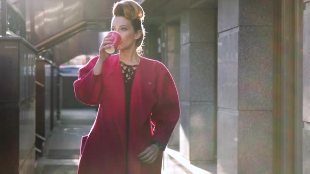 Ελκυστική νεαρή ξανθιά γυναίκα πίνει καφέ στις ακτίνες του ήλιου ρύθμιση. Κορίτσι στο φόντο της σύγχρονης αρχιτεκτονικής με καφέ. Αργή κίνηση — Αρχείο Βίντεο