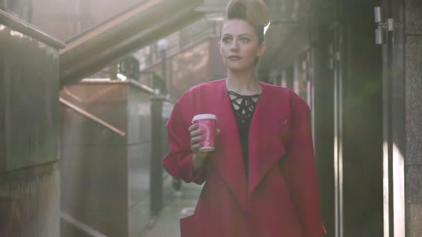 Confident woman with a beautiful unusual hairdress walks through the spring city. Girl on the background of modern architecture with coffee. SLOW MOTION — Stock Video