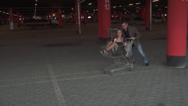 Killen rullar sin flickvän på en vagn i stormarknad parkeringen... unga människor som njuter utomhus med shopping vagn race. slowmotion — Stockvideo