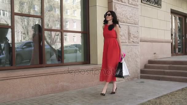 魅力的な女の子喜んで買い物袋が付いている通りを歩きます。赤いドレスの若い女性は、成功した買い物の後笑顔します。スローモーション — ストック動画