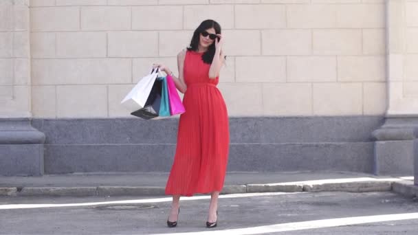 购物在一个有吸引力的女孩手中的包。模型鲜红色的裙子和太阳镜在镜头前摆姿势。女人微笑和欢乐。慢动作 — 图库视频影像