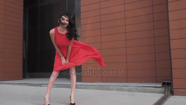 Sexy Mädchen im roten Kleid lächelt. Mädchen mit schönen Beinen in einem windgepeitschten Kleid. Wind kräuselt Gehrock. Zeitlupe — Stockvideo