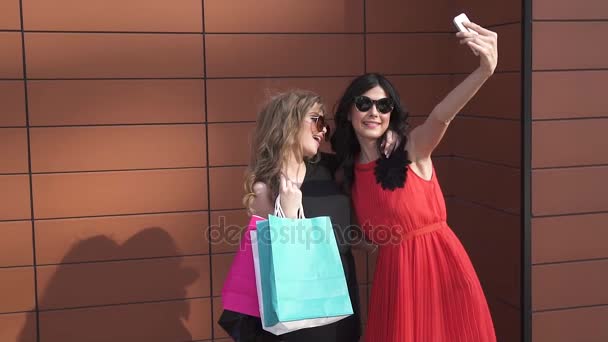 Twee vrienden maken een selfie met grappige gezichten. Zonnig portret van meisjes. mooie meisjes in zonnebril met shopping tassen in de hand nemen van foto's en lachend. Slow motion — Stockvideo