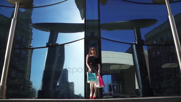 По магазинам. милая молодая девушка с сумками в руке в черном платье и солнцезащитные очки. блондинка на фоне современного здания из голубого стекла. Медленное движение — стоковое видео