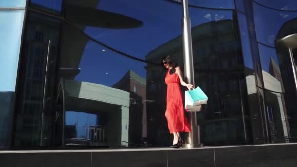 Mooi jong meisje in de rode jurk met shopping tassen in de hand genieten van het mooie weer. meisje wandelen langs de storefronts. Slow Motion — Stockvideo