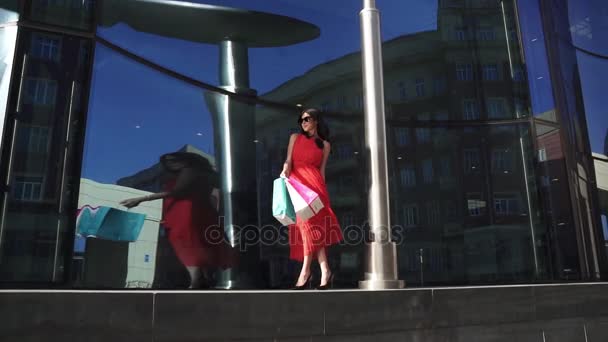 Menina bonita na montra da loja. A morena em um vestido vermelho e óculos de sol está segurando sacos de compras. Modelo europeu profissional posando na câmera. LOW MOTION — Vídeo de Stock