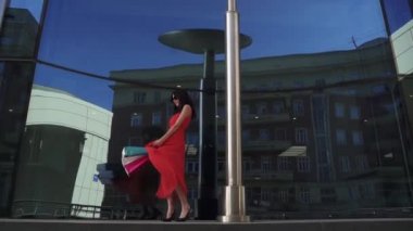 güzel model modern mimarisi fonunda poz elinde alışveriş torbaları ile. Kırmızı elbise ve güneş gözlüğü genç çekici kız. Ağır çekim