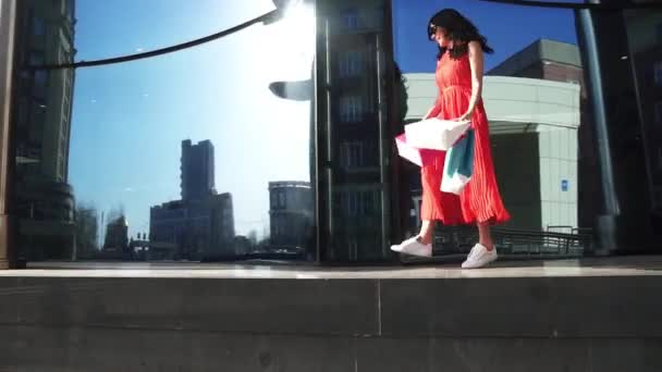 Красивая девушка в красном платье отражается в витринах магазина. Брюнетка с сумками для покупок в руках. Медленное движение — стоковое видео