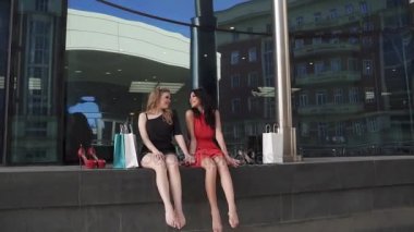 aktif alışveriş sonra en iyi arkadaşları mavi camdan yapılmış modern bir binada arka planı oturan bir dinlenme hakkına sahiptir. Yansımaları ve güneş ışınları. Kızlar beraber vakit ve gülümse. Ağır çekim