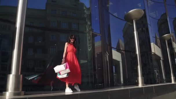 Παιχνιδιάρικο σέξι νεαρή κοπέλα σε ένα κόκκινο φόρεμα περπατώντας δίπλα σε ένα σύγχρονο κτήριο από μπλε γυαλί. Αντανακλάσεις και ηλιαχτίδες. Μελαχρινή με σακούλες για ψώνια στα χέρια. Αργή κίνηση — Αρχείο Βίντεο