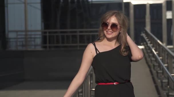 Vonzó lány egy rövid fekete ruhát és a napszemüveg. Profi modell pózol a kamera. lassú mozgás — Stock videók