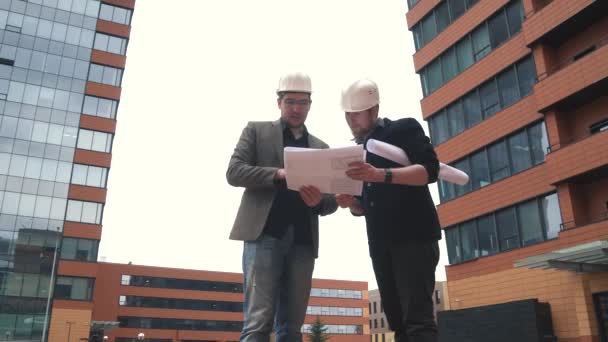 Polier und Architekt auf der Baustelle. Zwei Männer mit Bauhelmen auf dem Bauplatz, um den Plan zu besprechen, eine Geschäftsfrau bei der Arbeit zu bauen. Mann hält einen Entwurf in der Hand — Stockvideo